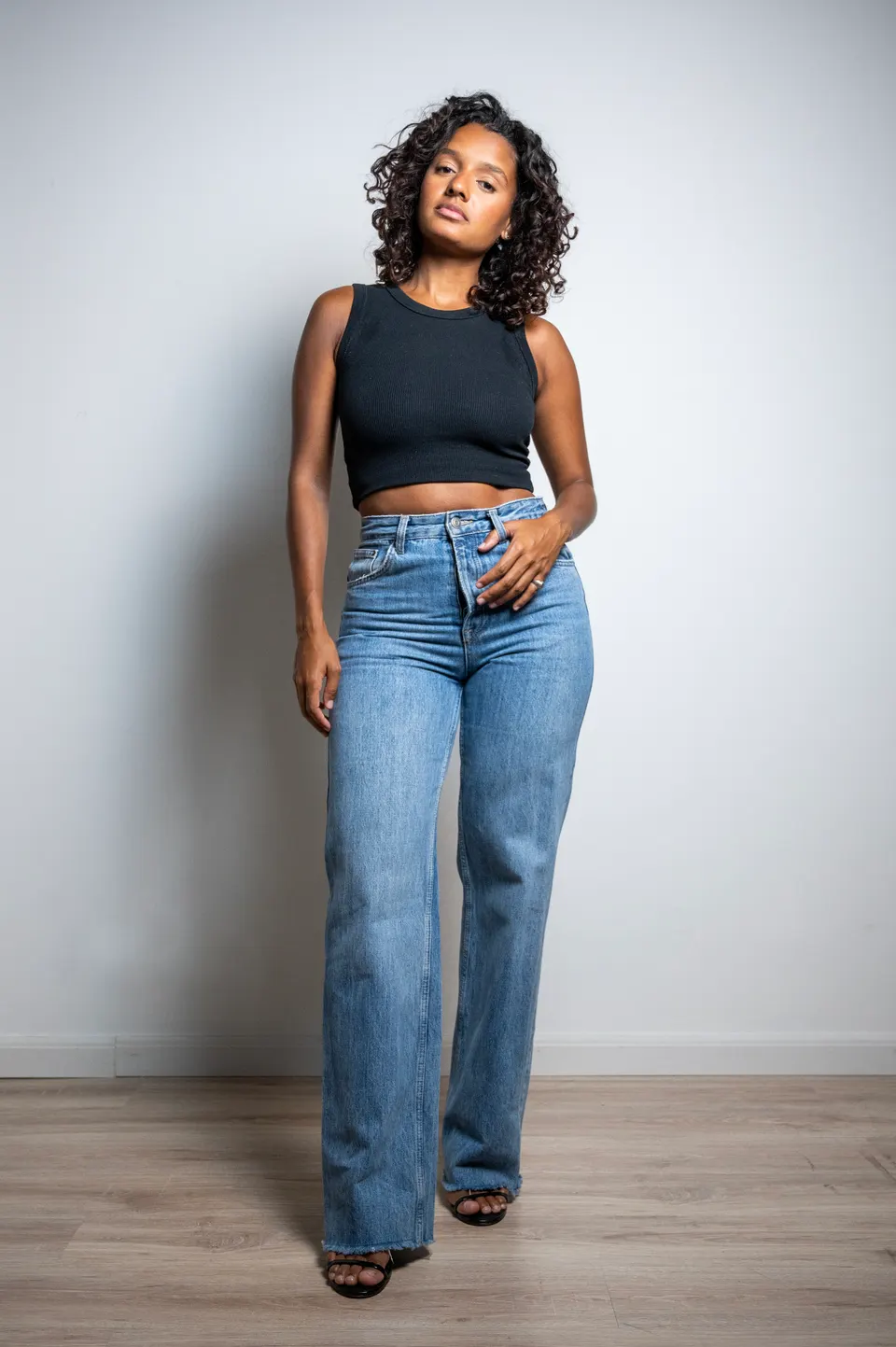 Mulher de cabelos cacheados, top preto e calça jeans, em pé diante de um fundo claro