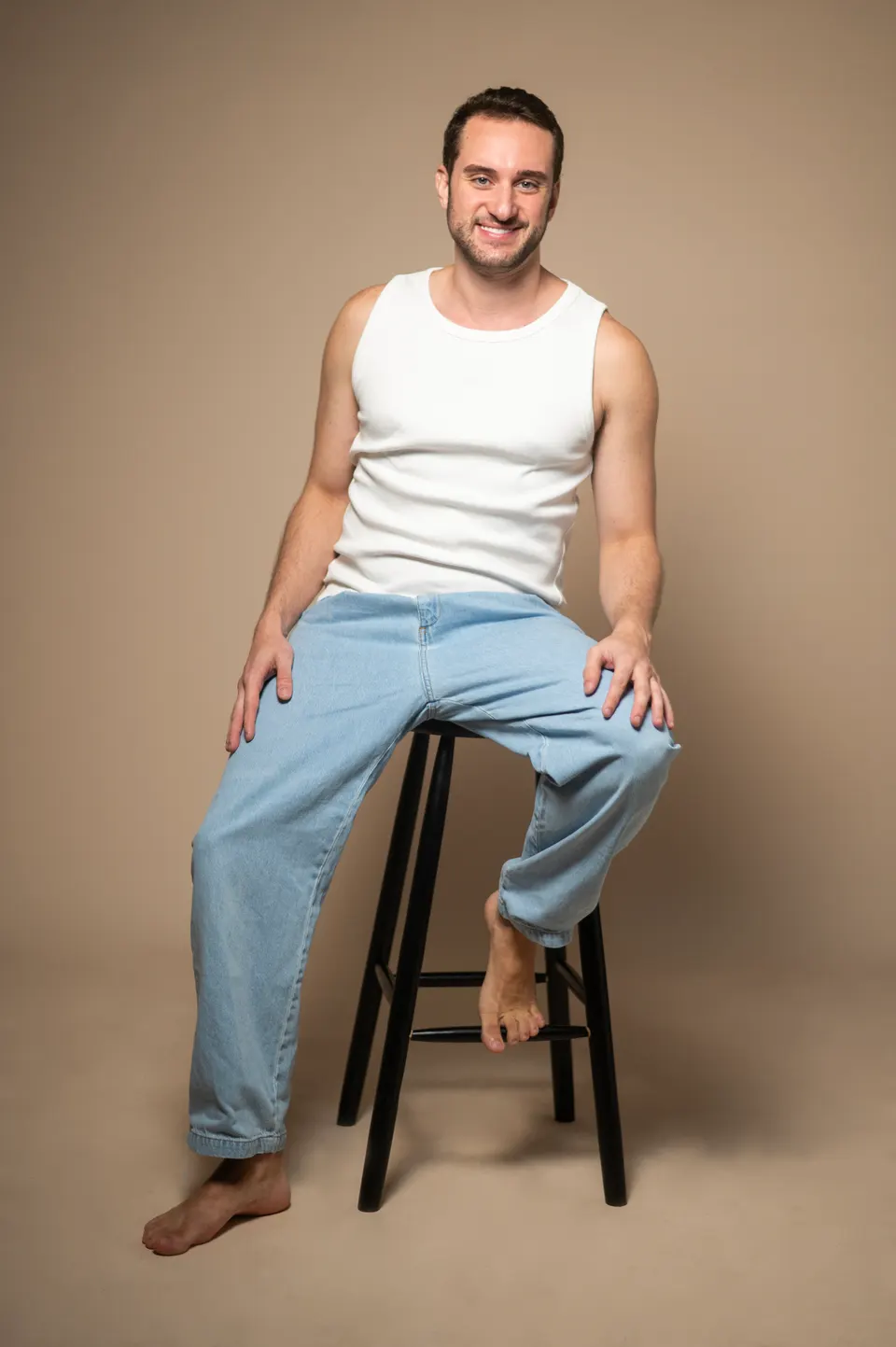 Homem de regata branca e calça jeans, sentado em um banco alto