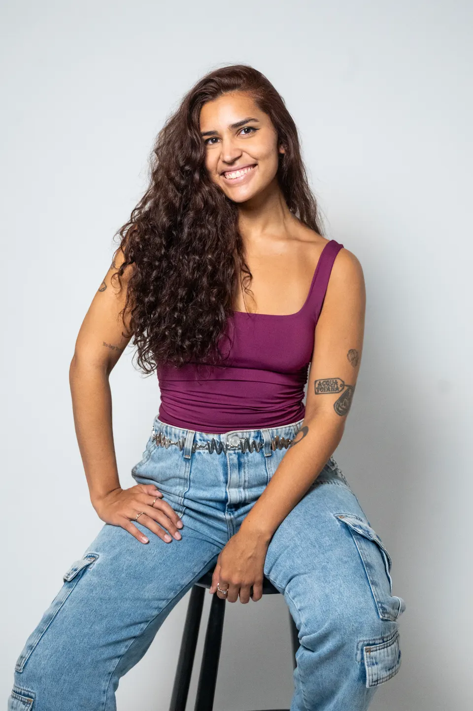 Mulher de cabelos cacheados longos e top vinho, sorrindo sentada em um banco
